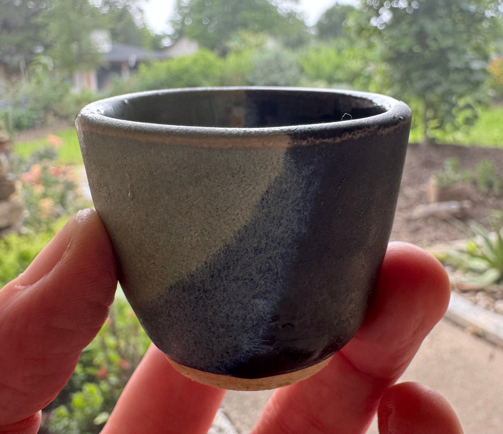 A handcrafted ceramic o-choko sake cup with a green and dark glaze, resting on a blue glass surface outdoors